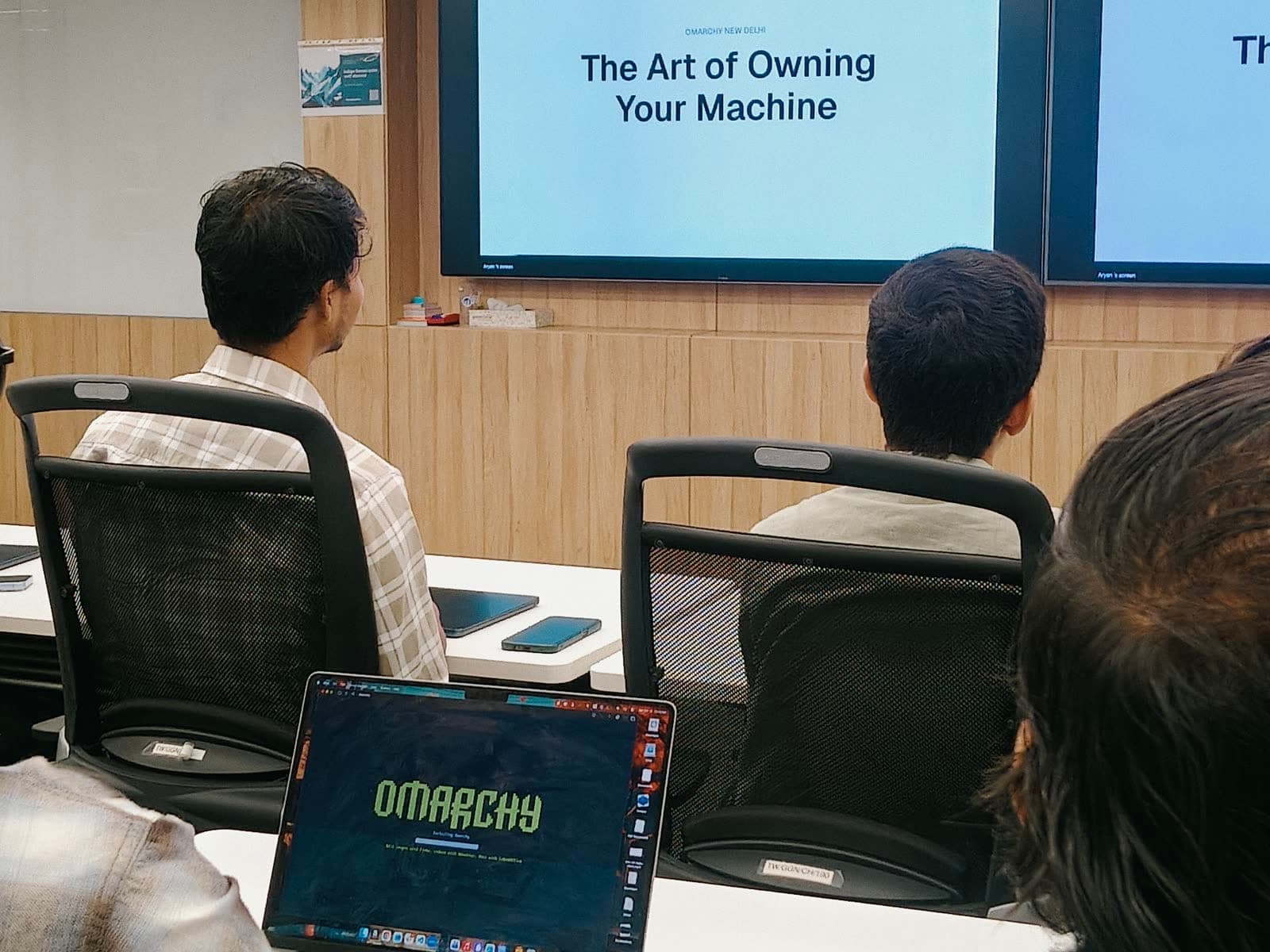 The Art of Owning Your Machine title slide on the big screens at Omarchy New Delhi, with an Omarchy laptop in the foreground